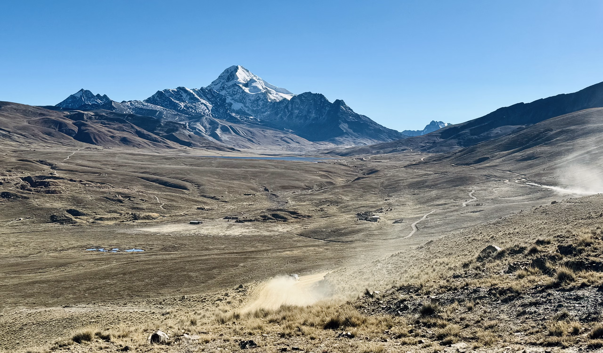 This breathtaking image captures the vast, rugged beauty of the Bolivian Andes, with the snow-capped peak of Huayna Potosí towering in the distance. The mountain’s sharp summit glistens under the clear blue sky, casting a majestic presence over the arid highland plain (Altiplano) below.