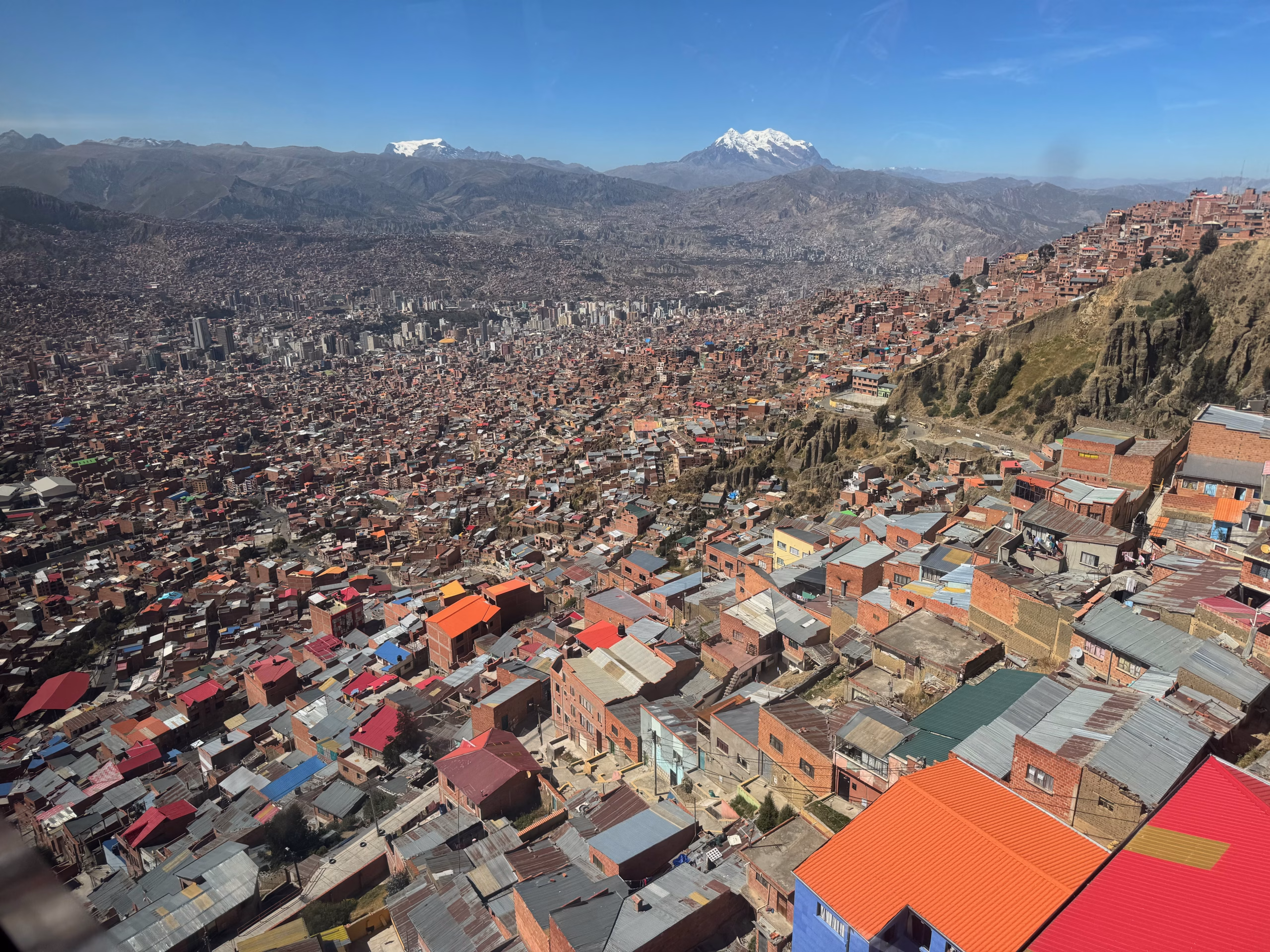A panoramic aerial view of La Paz, Bolivia, with colorful rooftops cascading down steep hillsides and Mount Illimani snowcapped in the background