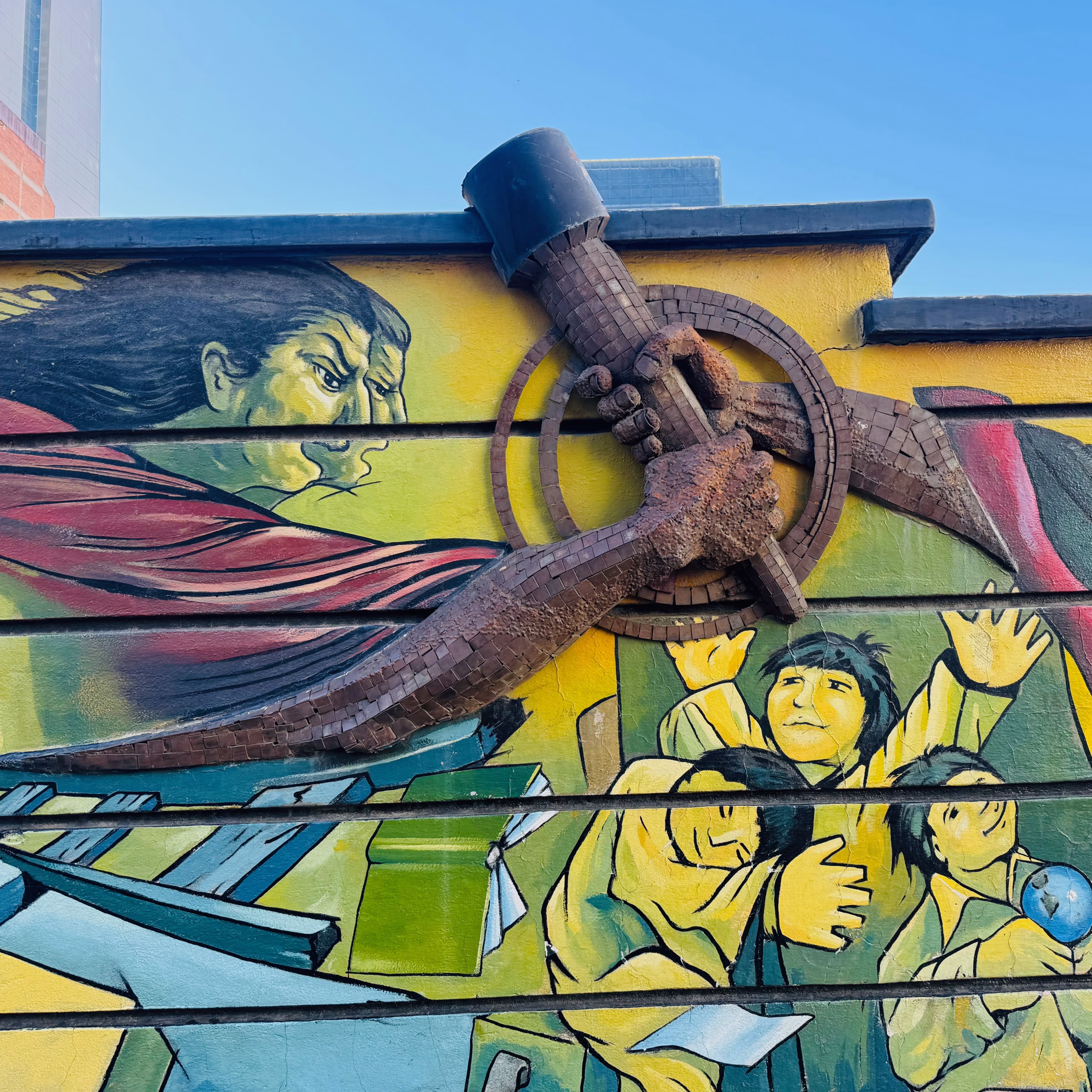 Colorful mural in La Paz, Bolivia, depicting a powerful arm holding a mining tool made of metal and tiles, symbolizing labor and resistance. The background shows expressive figures—an angry indigenous woman and distressed children—representing struggle, protest, and social injustice.