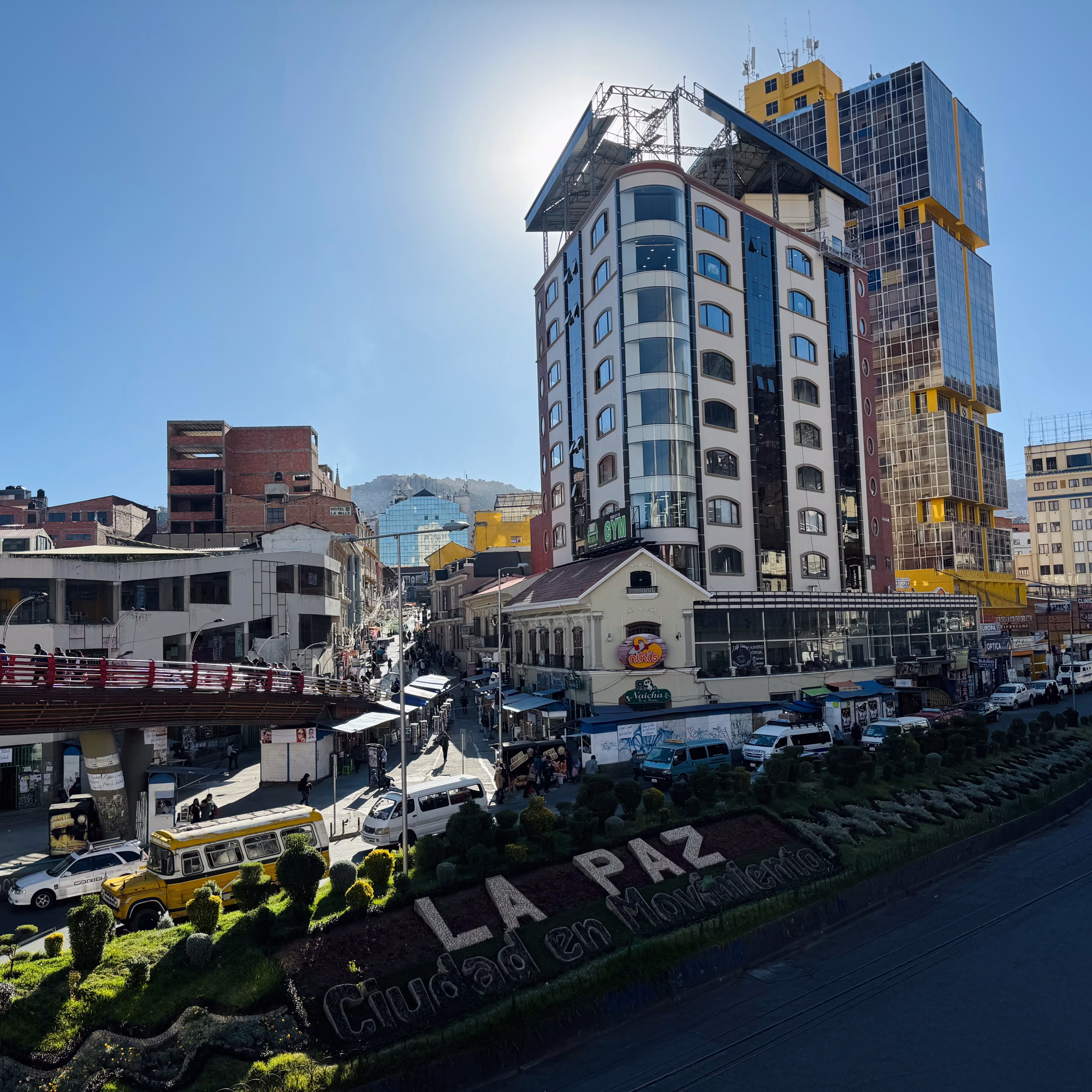 Cityscape of La Paz, Bolivia, showing a mix of modern high-rises and older buildings with a busy street scene below. A red pedestrian bridge crosses over traffic, and sculpted greenery spells out “La Paz Ciudad en Movimiento” in front.