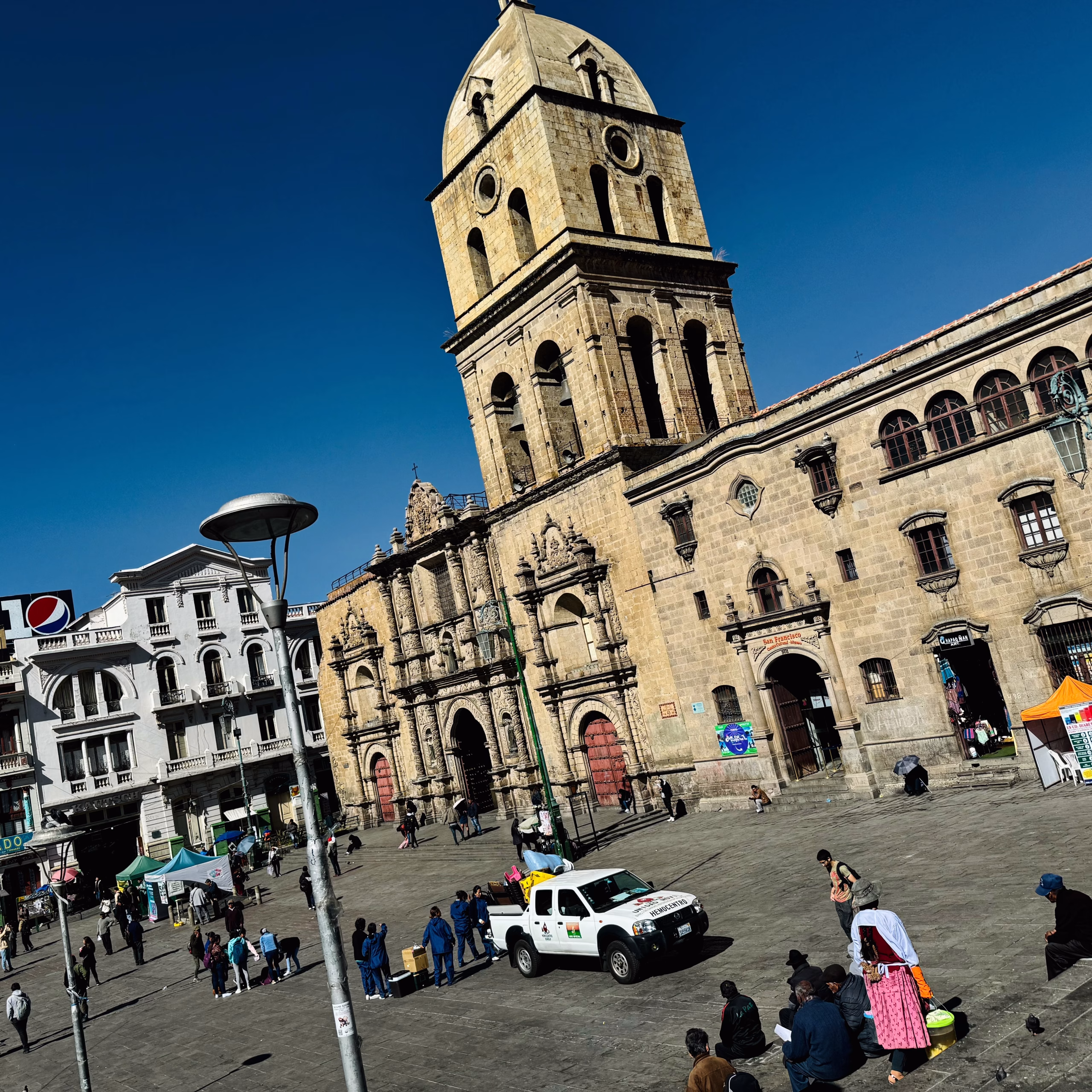 A view of the 16th-century San Francisco Church in La Paz, Bolivia, with its large stone bell tower and ornate baroque façade overlooking a busy public plaza filled with people, vendors, and a white city truck.