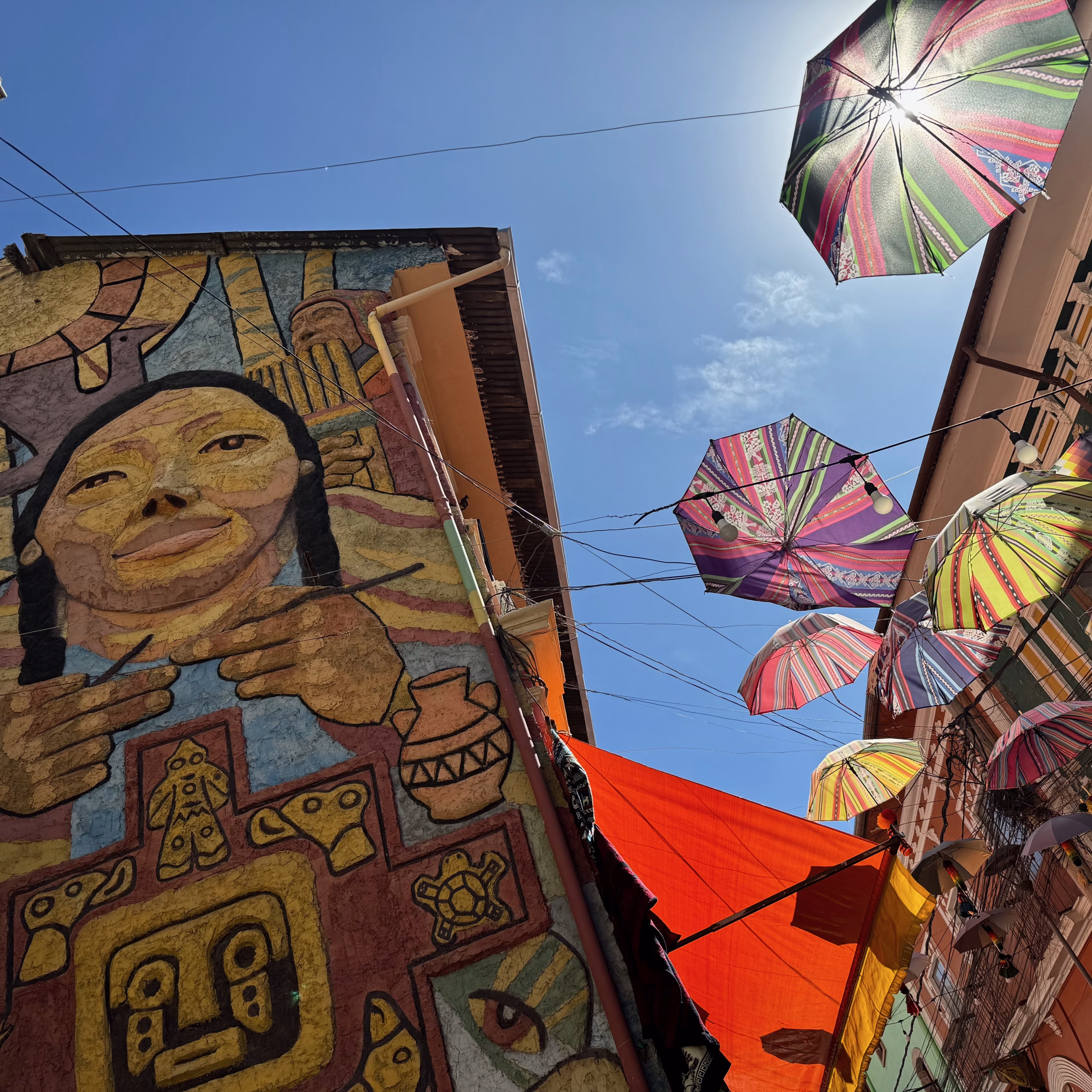 Vibrant mural of an indigenous woman surrounded by Andean symbols on a building wall in La Paz, Bolivia, with colorful umbrellas and cloths hanging overhead against a sunny blue sky.