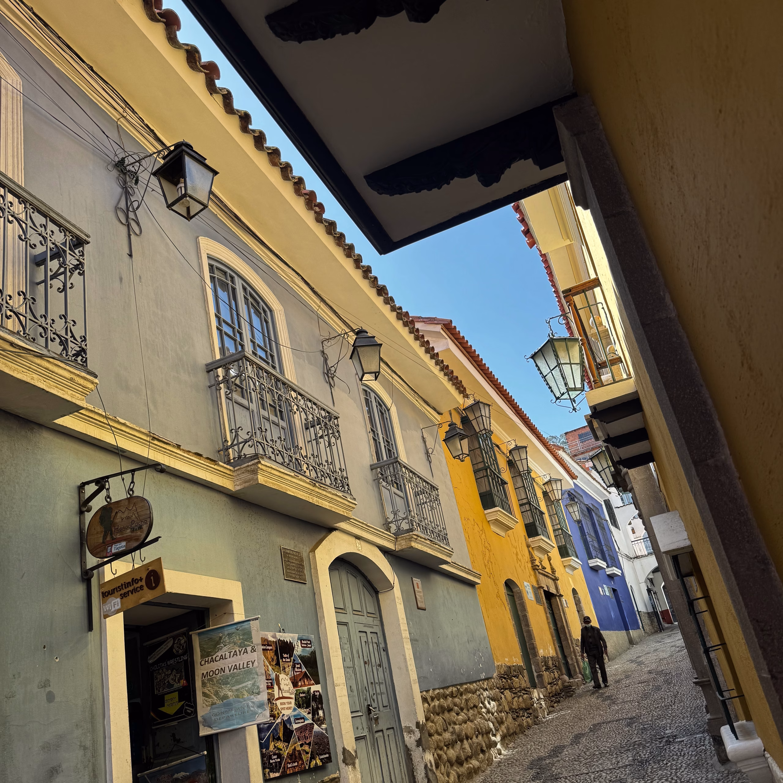 Narrow cobblestone street in La Paz, Bolivia, lined with colonial-style buildings painted in soft blue, yellow, and white, with wrought-iron balconies and vintage lanterns hanging along the walls.