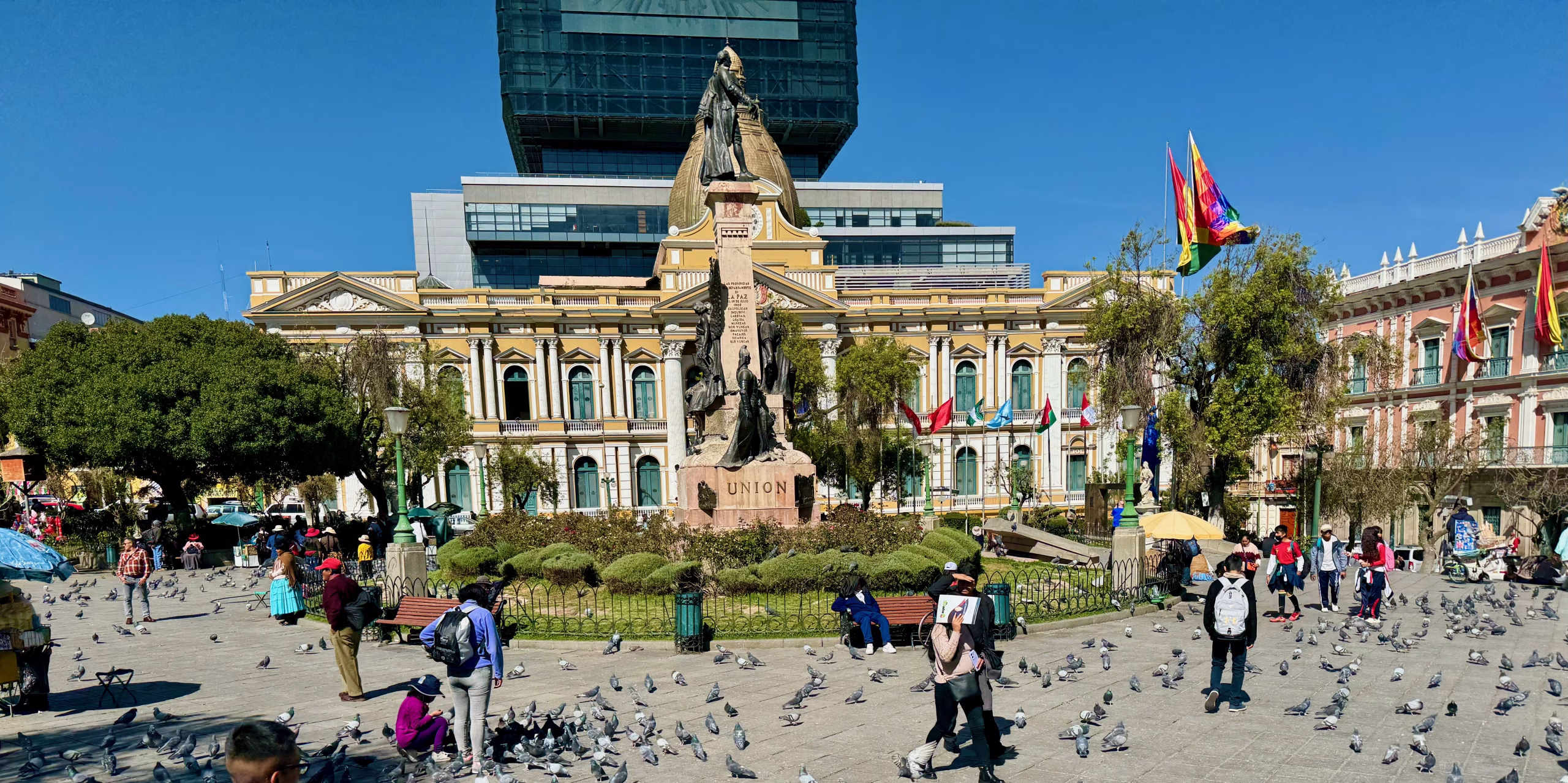 Wide view of Plaza Murillo in La Paz, Bolivia, with people walking and sitting among pigeons. At the center stands a large monument surrounded by trees and flowers, backed by historic government buildings and modern towers.