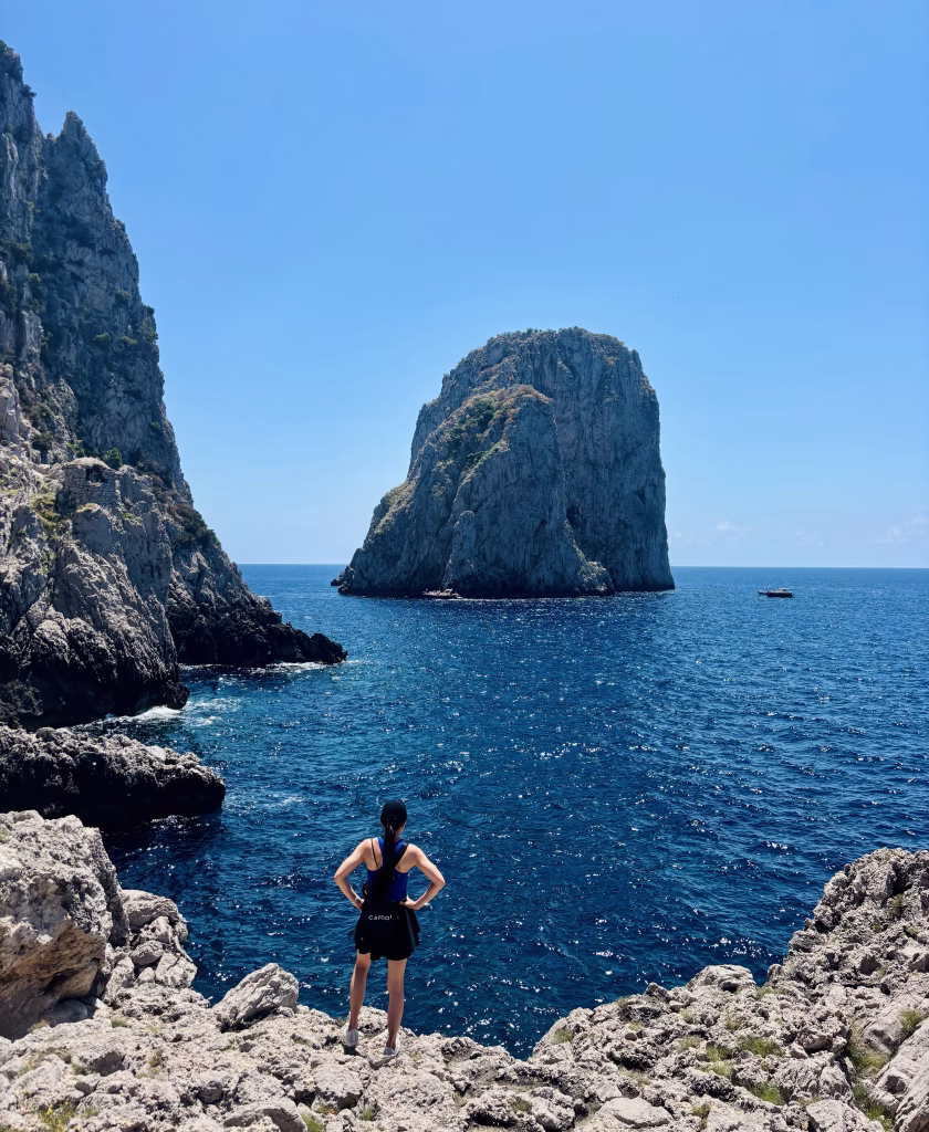 Kathy standing on rocky cliffs, looking out at the Faraglioni sea stacks rising from the deep blue waters of Capri.