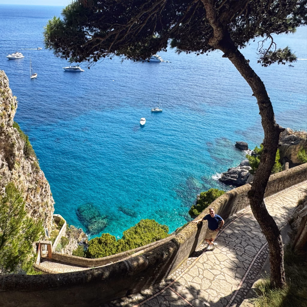 A scenic view of Via Krupp, the historic stone pathway that zigzags down a dramatic cliffside in Capri, Italy, framed by lush Mediterranean foliage and overlooking a serene turquoise bay dotted with anchored boats.
