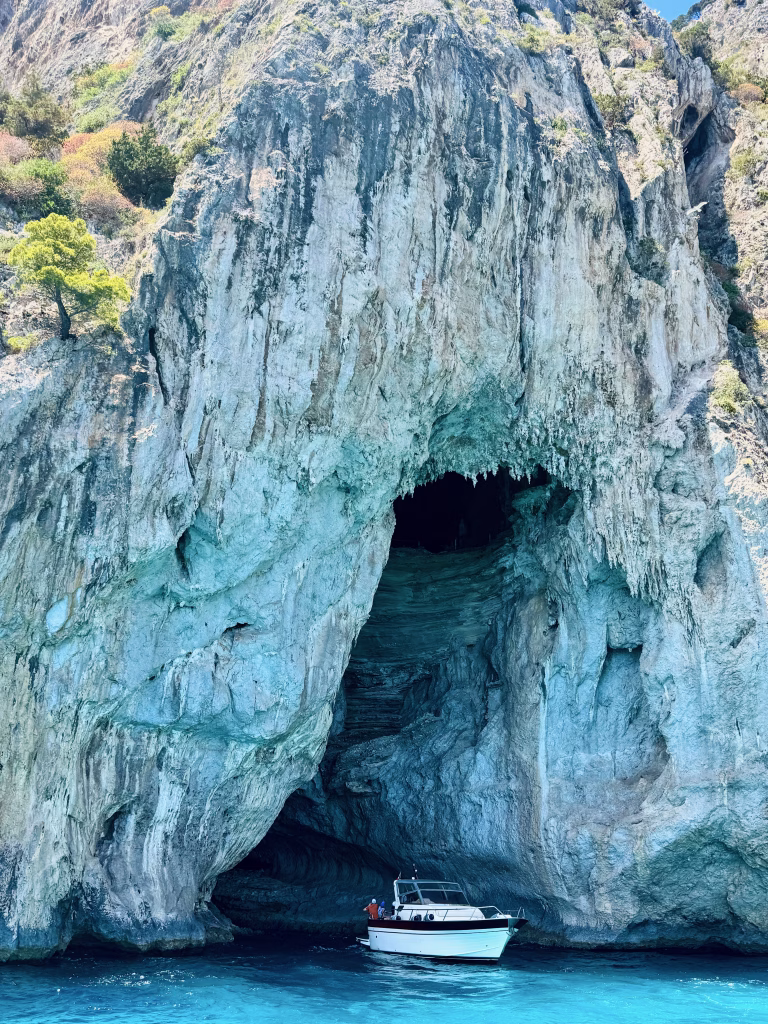 A small white boat enters Grotta Bianca (White Cave) on the coast of Capri, surrounded by towering limestone cliffs and turquoise blue water.