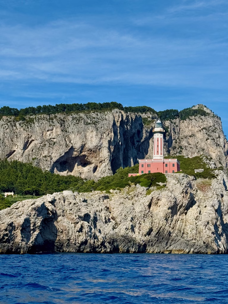 The pink and white Faro di Punta Carena lighthouse on the island of Capri, Italy, stands boldly atop rocky cliffs covered in greenery. Below, the clear blue sea laps against the rugged shoreline, while a bright sky with wispy clouds stretches overhead.