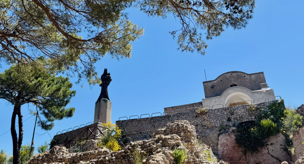 Statue of Madonna and Child and a small chapel at the top of Villa Jovis ruins in Capri, framed by pine trees and bright blue sky.