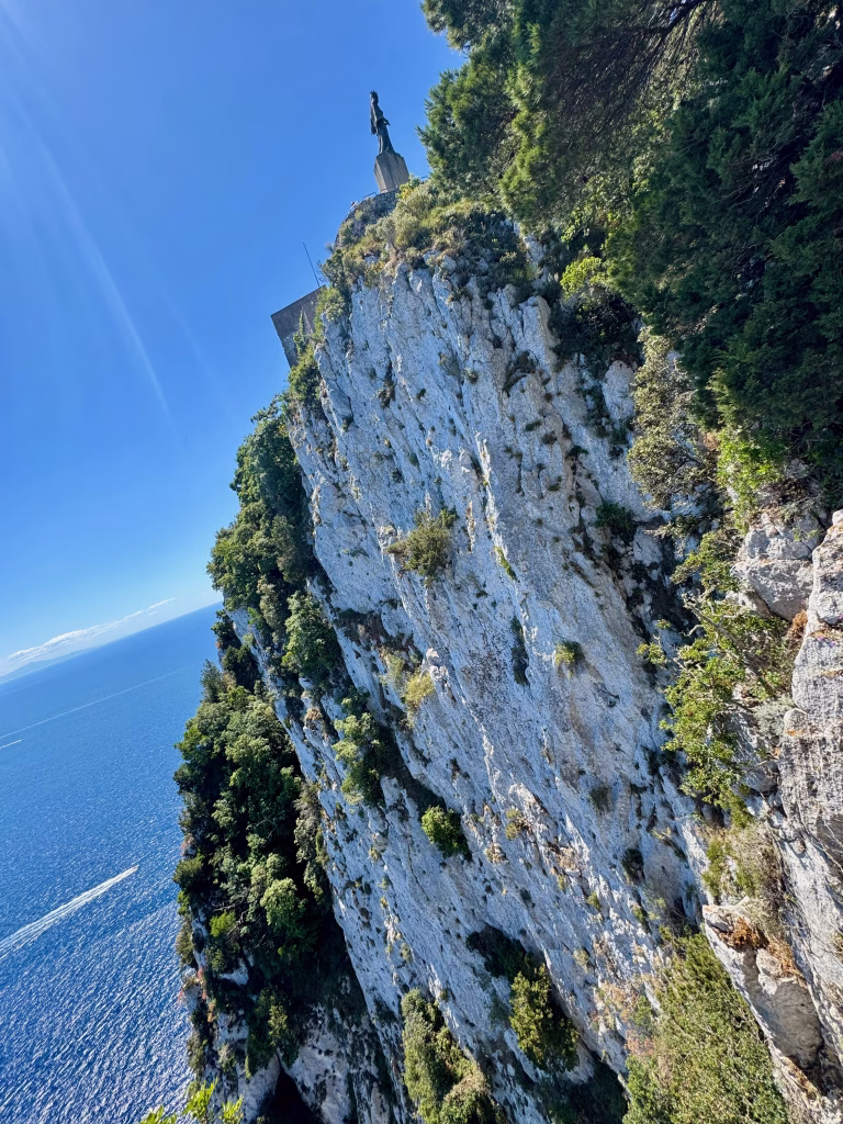 Statue atop a dramatic cliff at Villa Jovis on Capri, overlooking the deep blue sea, with green trees clinging to the rocky slope.