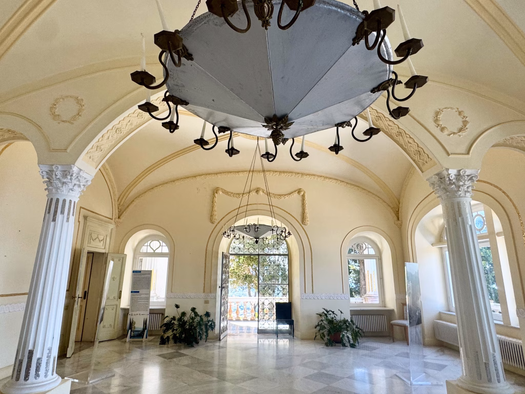 The grand main salon at Villa Lysis in Capri, Italy, featuring ornate neoclassical architecture. The space is adorned with tall white columns, intricate ceiling moldings, and a sparkling chandelier. Marble floors and elegant windows let in soft natural light, while decorative plants and an informational stand add character near a doorway leading outside.