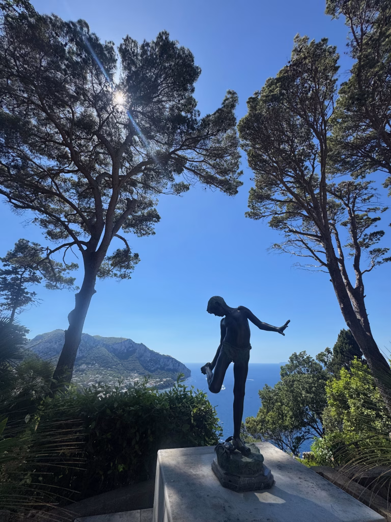 Bronze statue of a young fisherman adjusting his sandal at Villa Lysis in Capri, with the dramatic coastline and deep blue sea in the background.