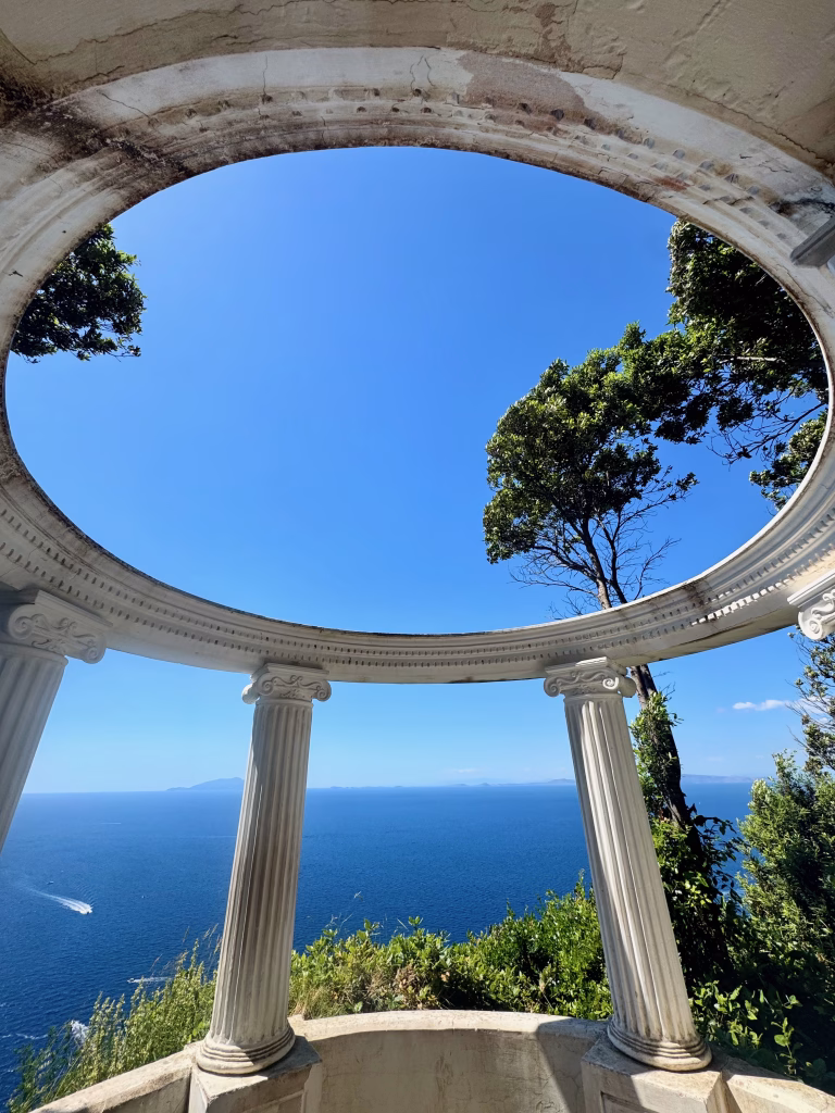 A neoclassical circular pavilion with ornate columns at Villa Lysis in Capri, Italy, frames a sweeping view of the deep blue sea and distant coastal cliffs. Mediterranean trees peek through the arches, and the sunlit sky stretches wide above, bathing the stonework in a warm glow.