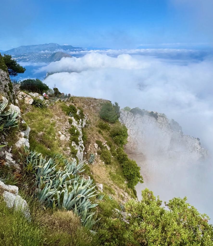 View from Monte Solaro with lush greenery and rocky cliffs fading into clouds, overlooking the Bay of Naples