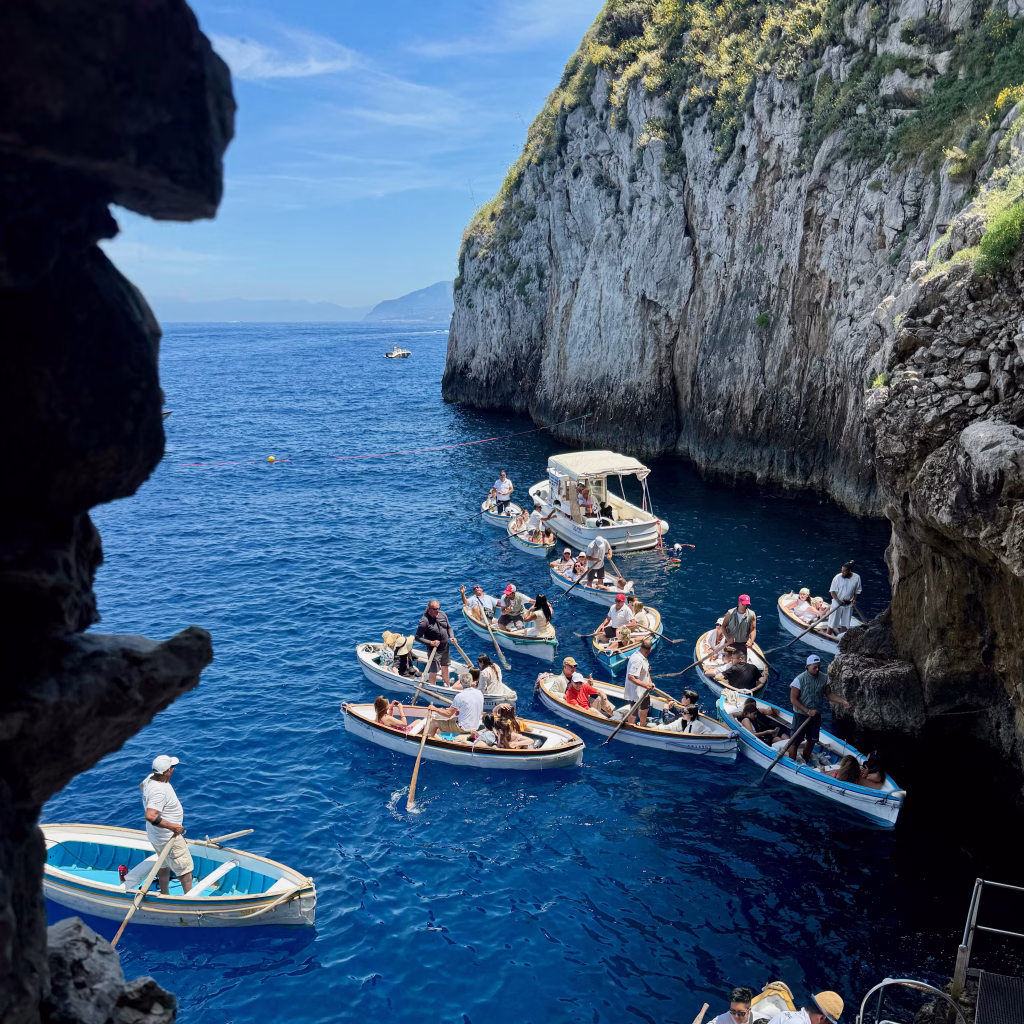 Cluster of small rowboats filled with tourists near the entrance of the Blue Grotto in Capri, with steep rocky cliffs rising beside the vivid blue sea