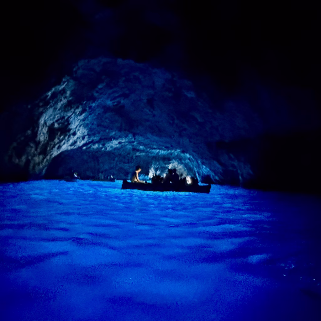 A small rowboat glides through the Blue Grotto, a sea cave off the coast of Capri, Italy. Inside, the cavern walls are bathed in an ethereal electric blue glow, created by sunlight filtering through underwater openings. Silhouetted passengers marvel at the otherworldly radiance dancing across the shimmering water.