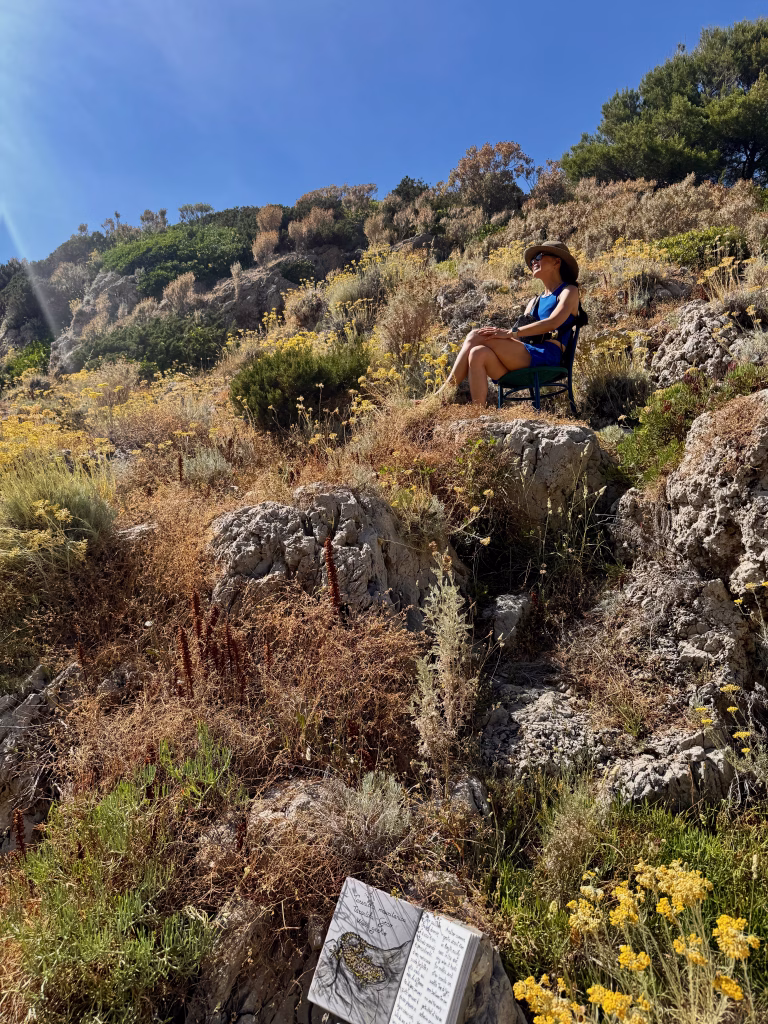 Kathy sitting on a chair in the sun-drenched hills along the Path of Forts in Capri, surrounded by wildflowers and rocky terrain, with a ceramic book in the foreground