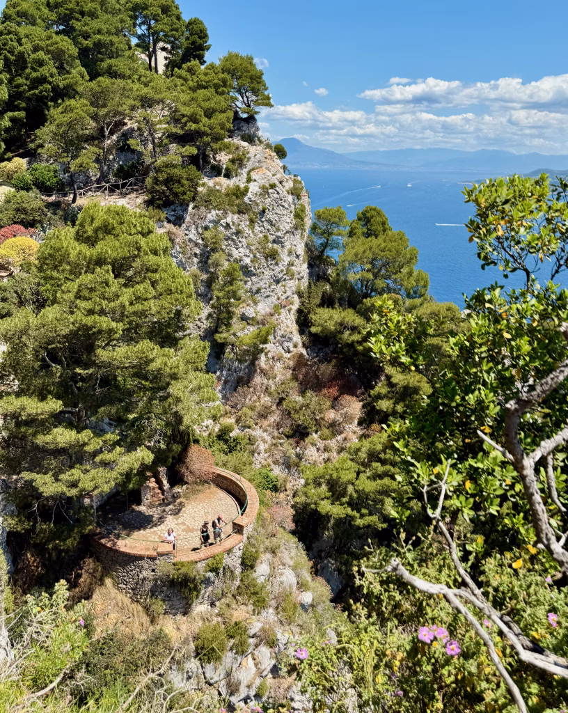 Coastal view from Parco Astarita showing lush cliffs, stone terraces, and boats on deep blue waters with the Sorrentine Peninsula in the distance.