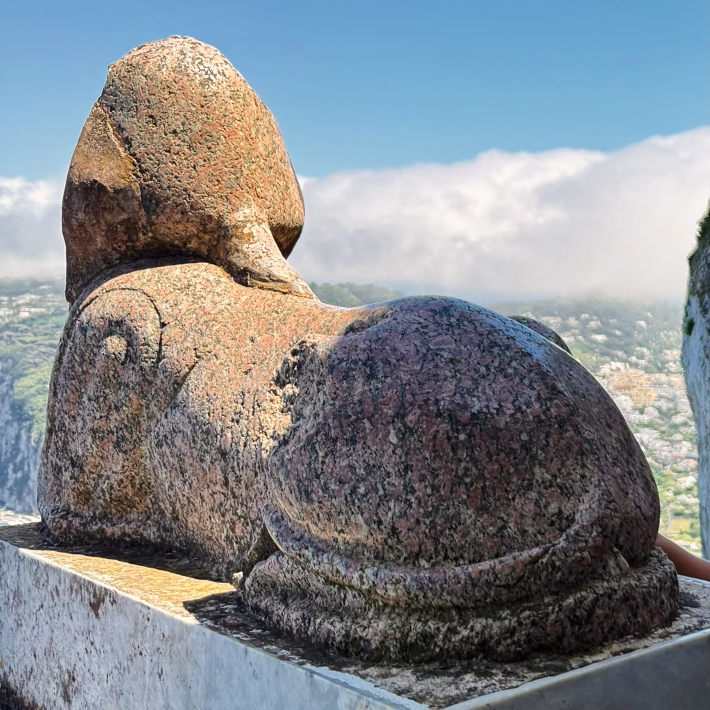 Rear view of the ancient granite sphinx statue at Villa San Michele in Anacapri, perched on a ledge with clouds and townscape in the background
