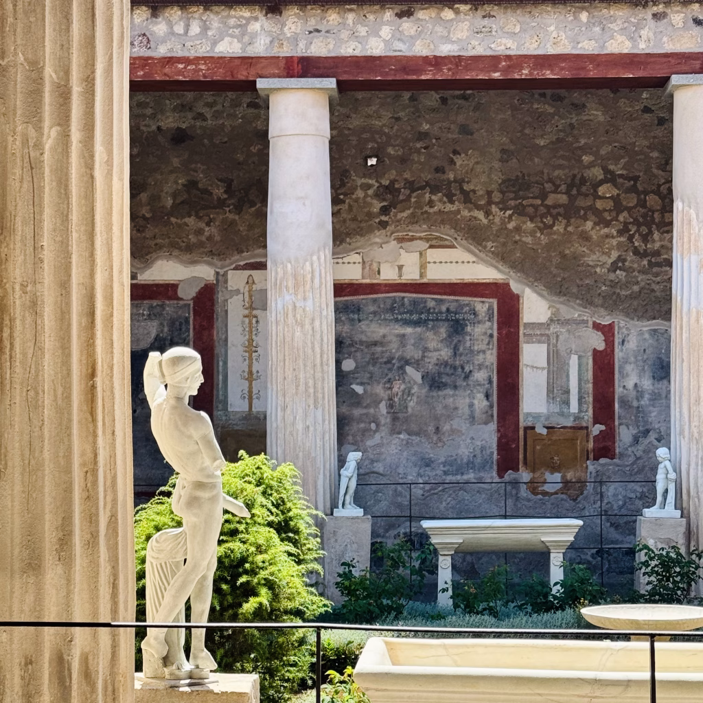 A well-preserved courtyard in Pompeii featuring marble statues, columns, and a painted fresco wall with red borders and classical motifs.