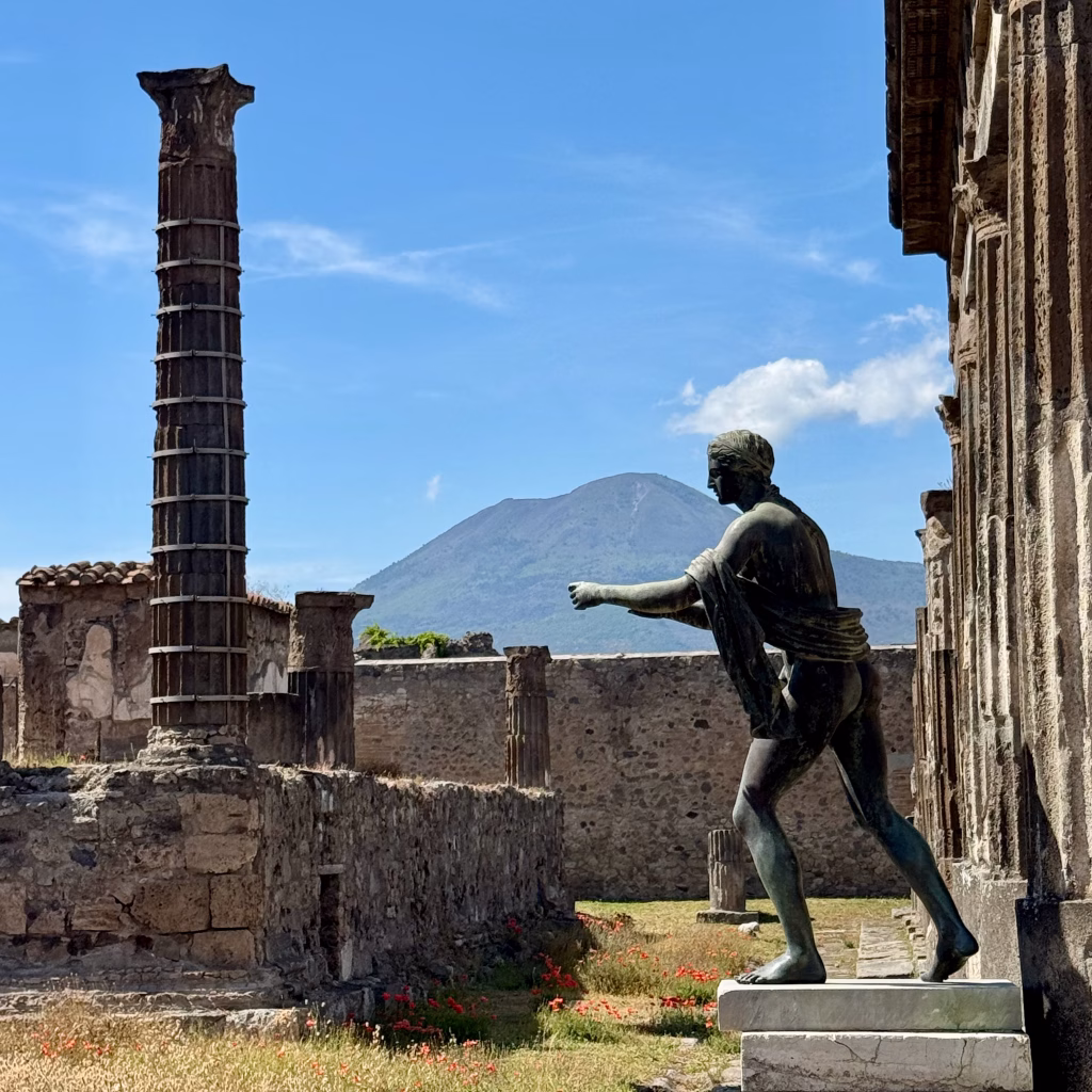 A bronze Roman statue stands in a ruined temple in Pompeii, with Mount Vesuvius rising in the background under a blue sky.