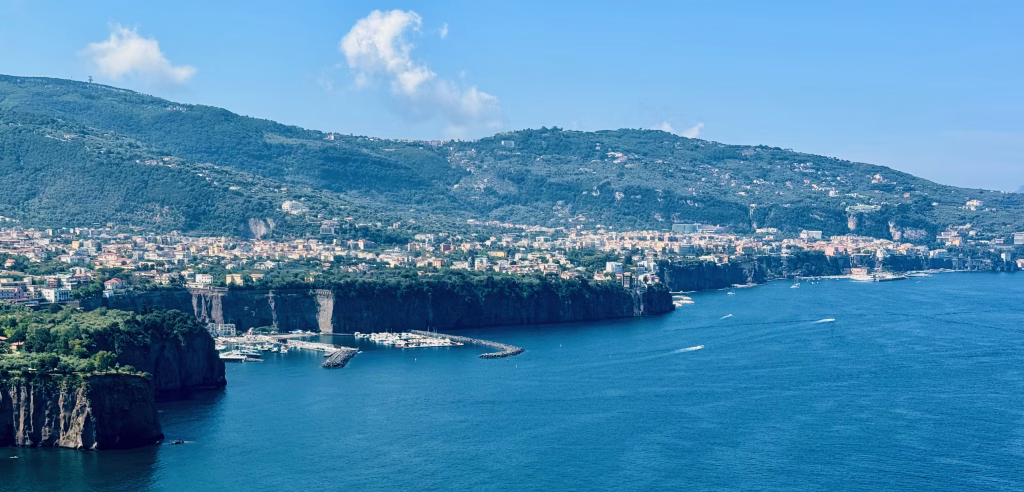 A sweeping coastal view near Sorrento, Italy, showcasing cliffside buildings and terraced gardens overlooking the vibrant Tyrrhenian Sea. A marina lined with boats rests at the base of the cliffs, while verdant hills rise in the distance under a bright blue sky streaked with wispy clouds.