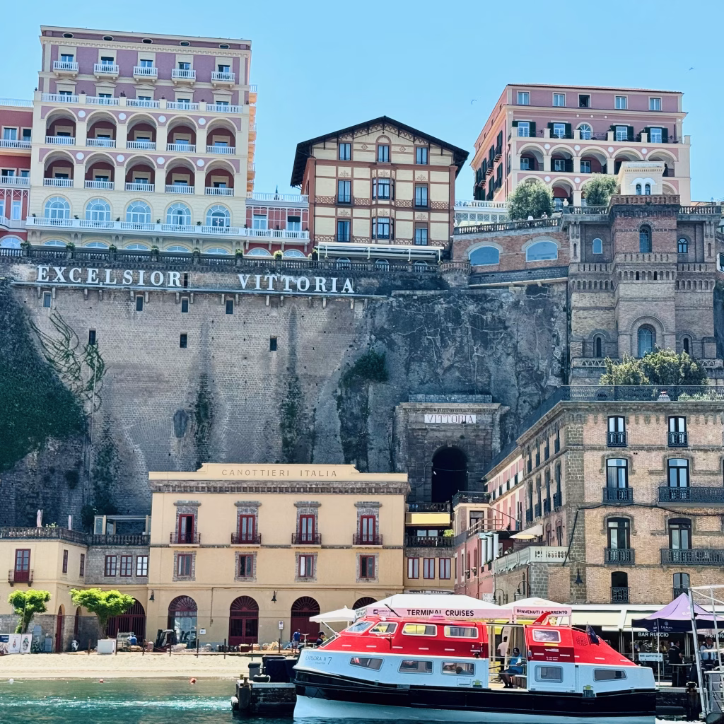 A striking coastal scene in Sorrento, Italy, where elegant buildings—including the grand Hotel Excelsior Vittoria—are perched dramatically atop a cliff. Below, a dock features the Canottieri Italia building and a red-and-white "Terminal Cruises" boat moored at the waterfront. The layered architecture and lush greenery blend seamlessly into the natural rock formation, framed by clear skies and calm sea.