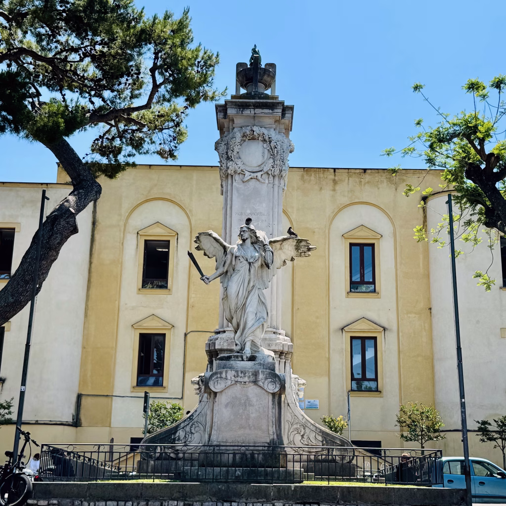 A majestic statue of a winged figure holding a sword and laurel wreath towers over Piazza della Vittoria in Sorrento, Italy. Set against a bright yellow building with arched windows and surrounded by leafy trees, the monument exudes classical grandeur. A bicycle rests nearby, adding a quiet note of everyday life beneath the clear blue sky.