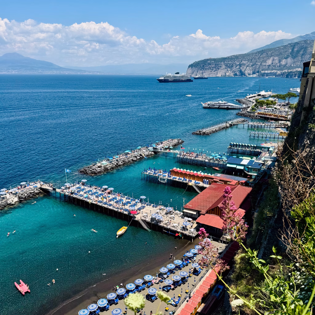 A vibrant coastal beach scene near Sorrento, Italy, featuring multiple wooden piers stretching over turquoise waters. Each pier is lined with blue sun loungers and umbrellas, while swimmers enjoy the calm sea below. The shoreline is backed by steep, rocky cliffs covered in greenery, with boats and a cruise ship visible in the distance.