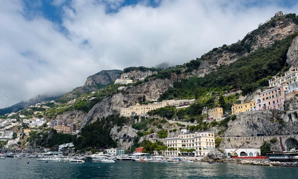Colorful buildings and dramatic cliffs of Amalfi town viewed from the sea under a cloudy sky.