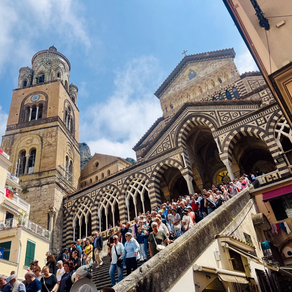 Crowds descend the grand staircase of Amalfi Cathedral, framed by its striped Romanesque façade and bell tower under a partly cloudy sky.