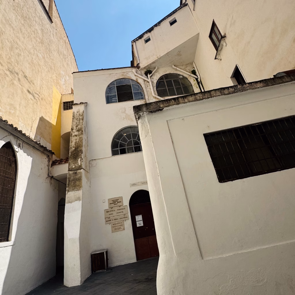 A historic alleyway in Amalfi, Italy, featuring whitewashed and beige buildings with arched windows, a weathered stone pillar, and signage identifying cultural institutions including the Cappella di Santa Maria, Biblioteca Comunale, and Archivio Storico Comunale. Bright sunlight and clear blue sky frame the scene, highlighting architectural details and aged textures.