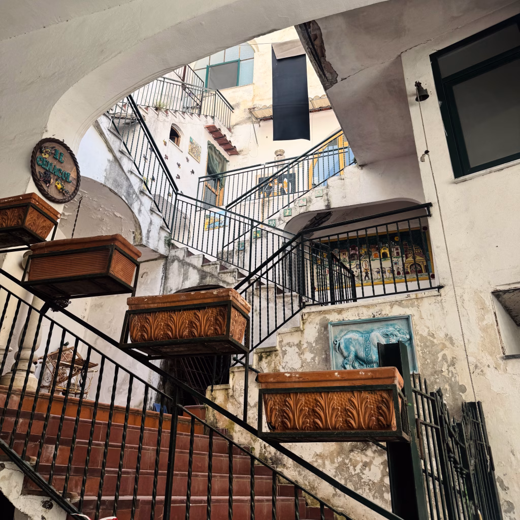 A colorful multi-level staircase in Amalfi, Italy, featuring red tile steps with black metal railings adorned with wooden planters. The surrounding walls are aged and textured, decorated with eclectic art—including a blue ceramic cow relief, vibrant murals, and a round sign reading “Le Chiacchiere.” Multiple balconies and windows suggest a historical setting rich in character and charm.