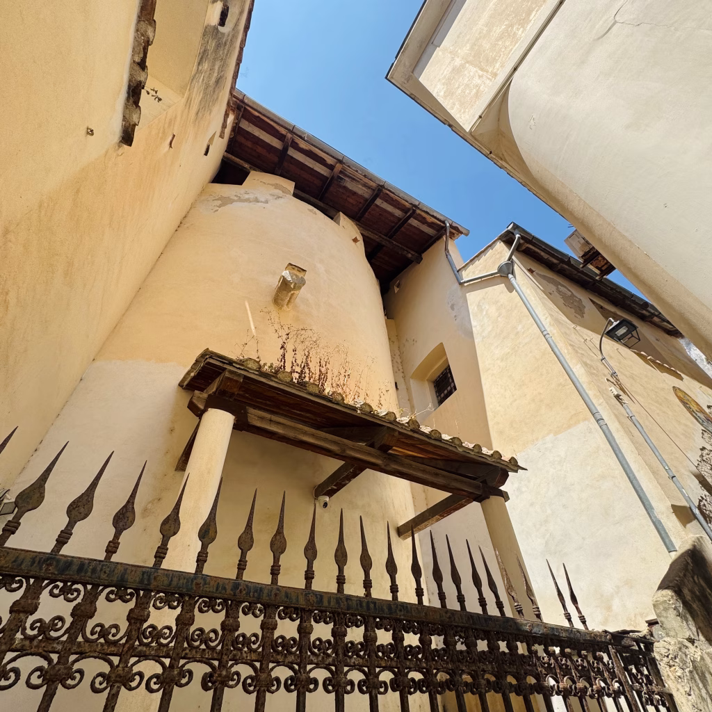 A historic corner of a beige building in Amalfi, Italy, featuring a small balcony-like structure with a wooden roof supported by white columns. A pointed metal fence runs along the foreground, while a drainpipe and vintage-style streetlight adorn the wall. The clear blue sky and rustic architecture evoke a quiet Mediterranean elegance.