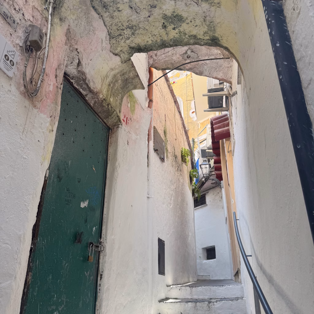 A narrow stone alleyway in Amalfi, Italy, with worn white walls and moss-covered archways framing a green wooden door marked with graffiti and a padlock. The staircase ascends between historic buildings adorned with small balconies, exposed wires, and aged textures, creating a sense of quiet, lived-in charm.
