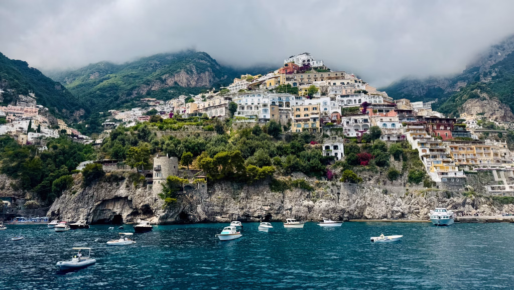 Colorful buildings of Positano cascade down steep cliffs toward the sea, with boats floating in the blue water and clouds hugging the mountains above.