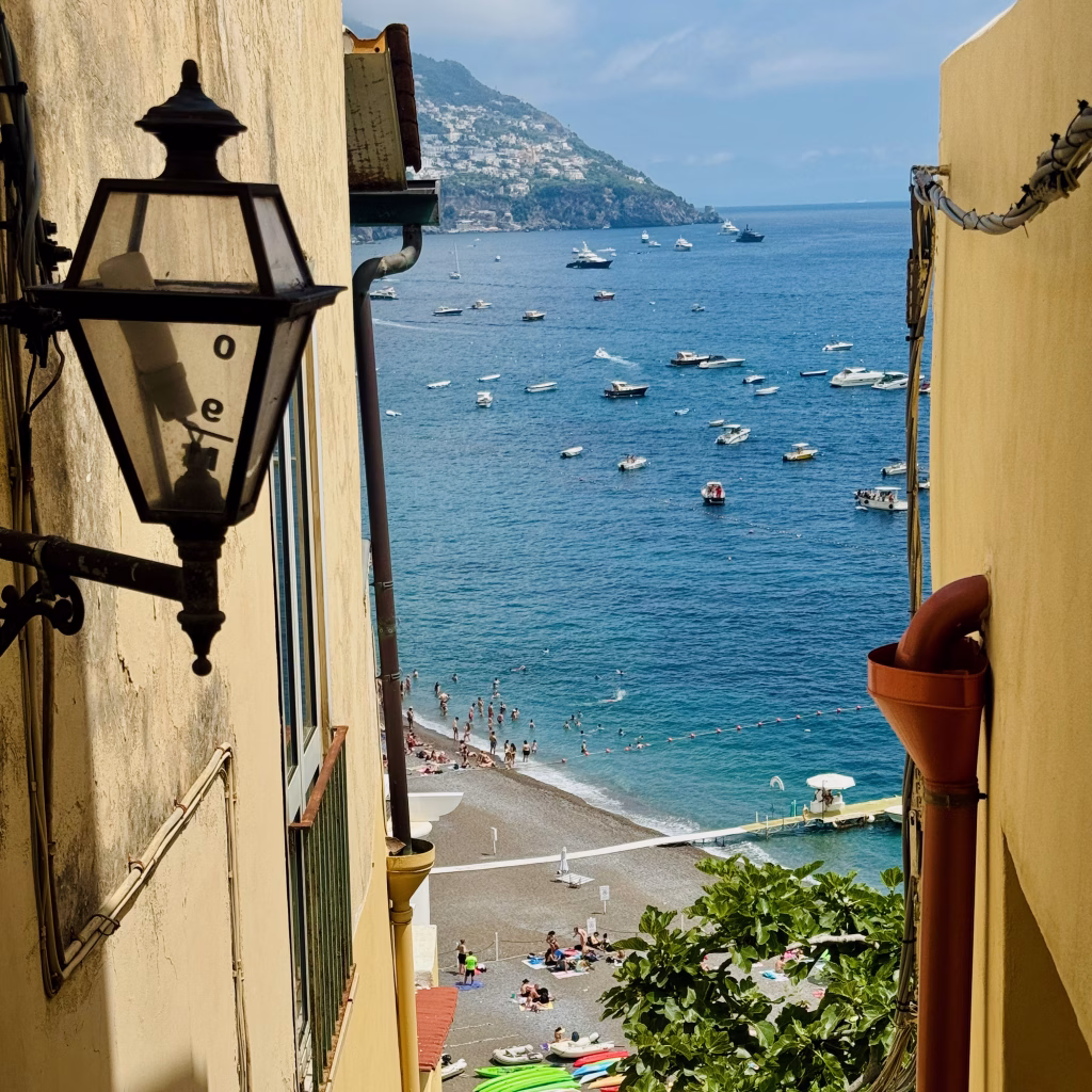 Narrow alleyway with a vintage lantern leading to a beach below, with boats dotting the blue waters of the Positano coastline.