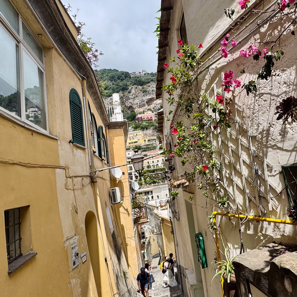 A charming stone alleyway in Positano, Italy, flanked by vibrant buildings with green shutters, flowering vines, and cascading blooms. The narrow path descends in steps toward distant views of the coastline and sea, capturing the town’s layered architecture and Mediterranean ambiance.
