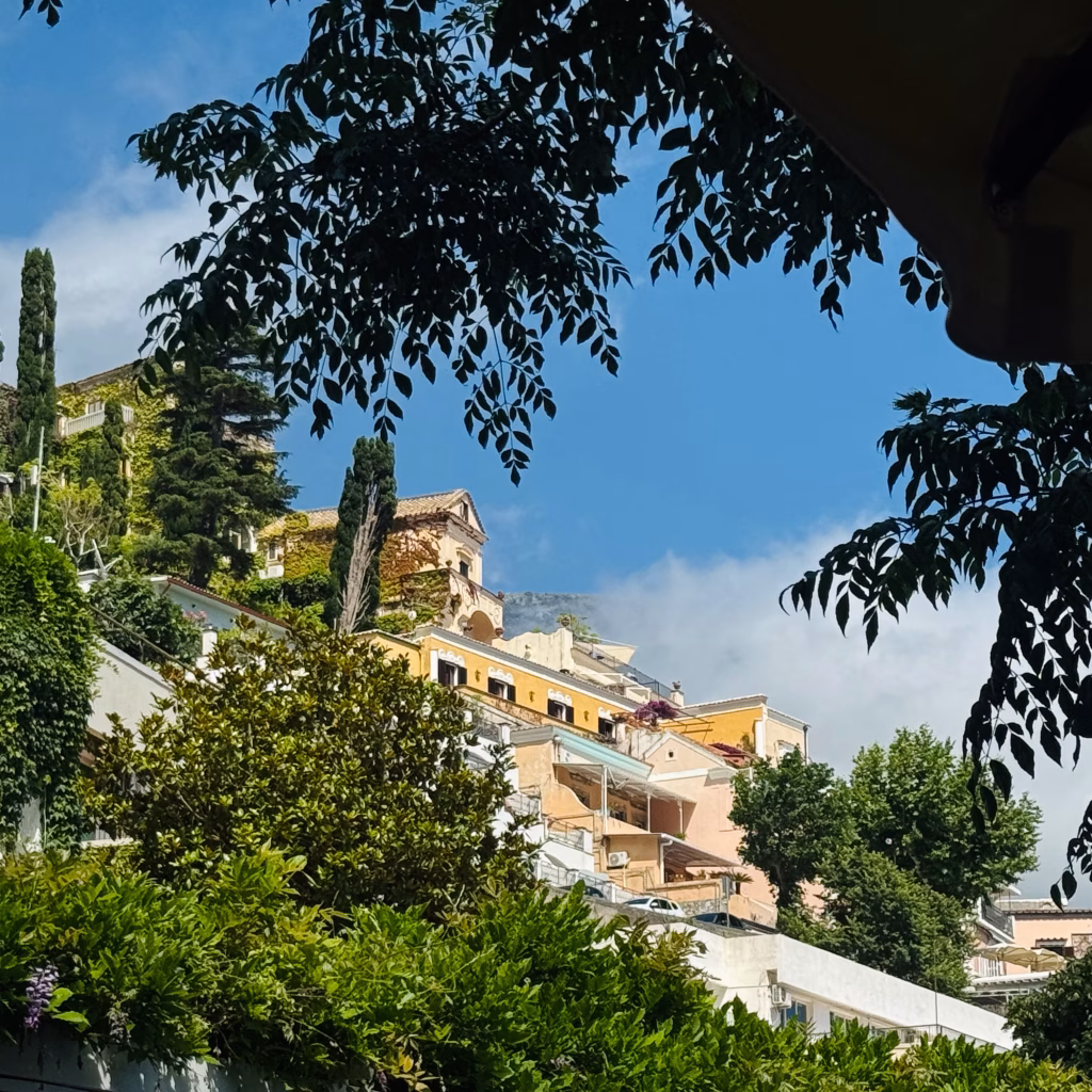 Colorful hillside buildings in Positano peeking through lush green trees under a blue sky.
