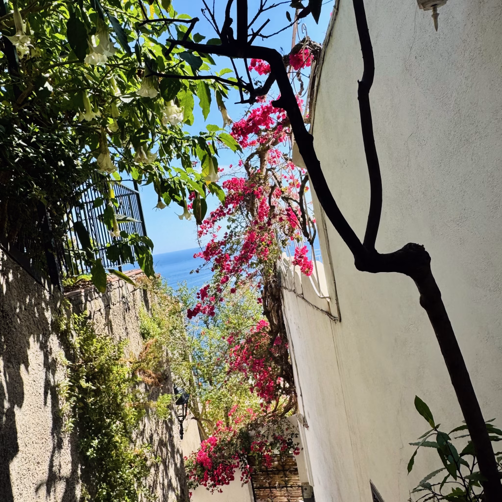 A sunlit coastal alley in Positano, Italy, featuring whitewashed and stone walls adorned with lush greenery and vibrant pink bougainvillea. The path opens toward a distant view of the shimmering Tyrrhenian Sea, partially framed by flowering vines and shadows cast by the bright summer sun.