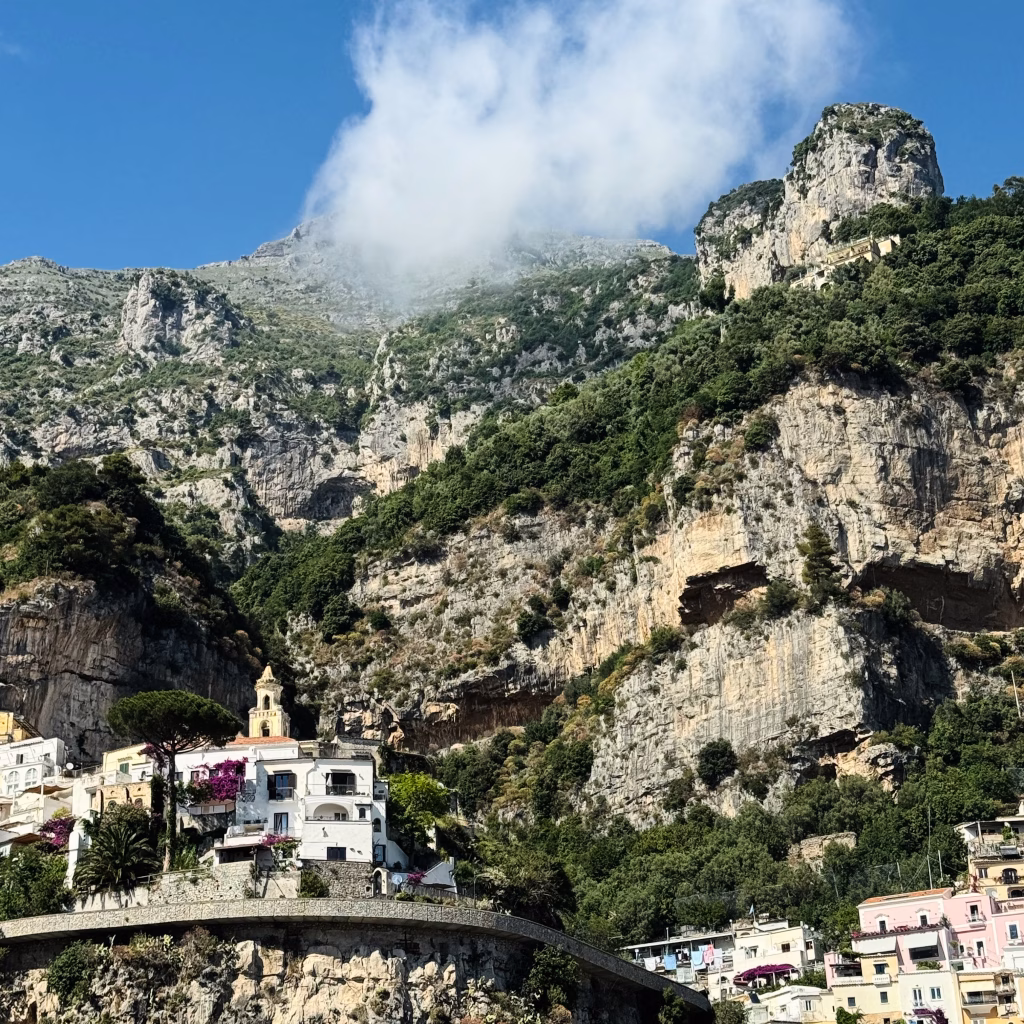 scenic view of a coastal town nestled at the base of towering cliffs covered in lush greenery. A prominent white building with a bell tower sits among smaller structures surrounded by colorful flowers and trees. The cliffs rise dramatically into a clear sky dotted with light clouds.