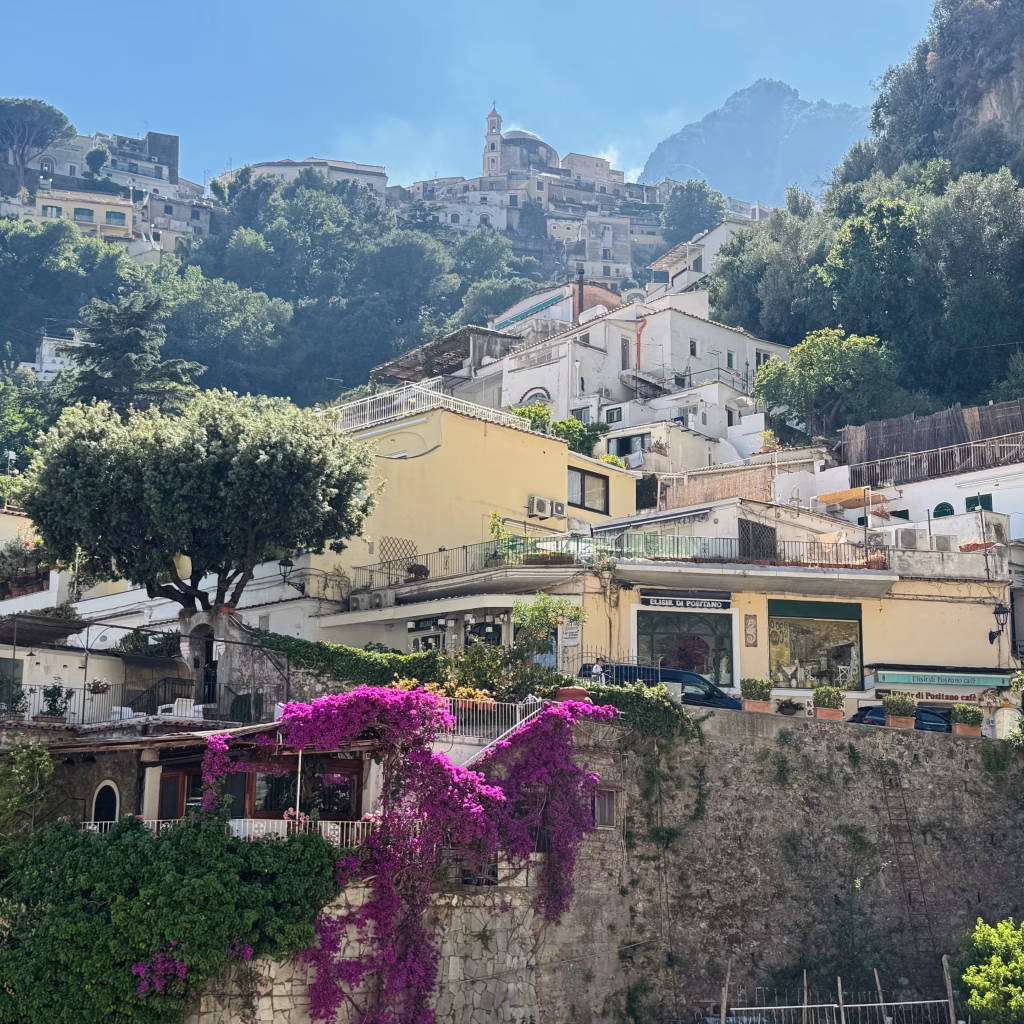 A sun-drenched hillside part of Positano on the Amalfi Coast, featuring layered buildings in soft earth tones and terracotta roofs rising toward a prominent cathedral with a domed bell tower. In the foreground, vibrant purple bougainvillea spill over a rustic stone wall, adding color and contrast to the lush greenery that weaves through the landscape.