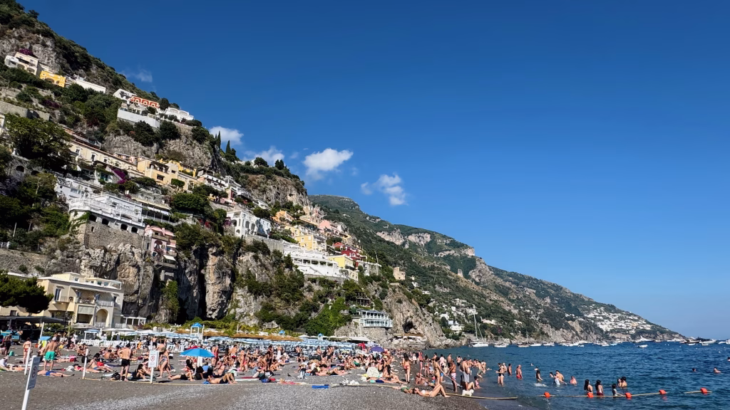 Crowds relax and swim on the main beach in Positano, with colorful cliffside buildings rising behind and boats dotting the blue Mediterranean Sea.